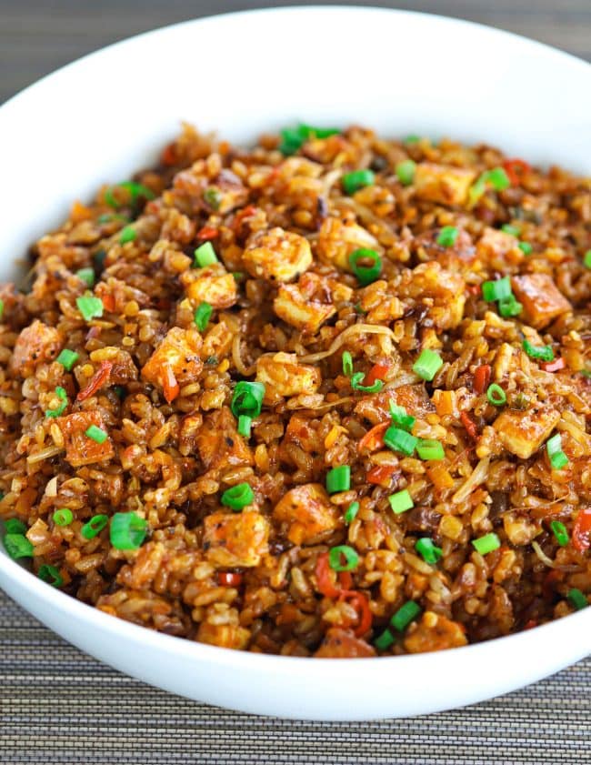 Front view of a white serving bowl filled with Crispy Tofu Spicy Fried Rice and garnished with chopped spring onion on a brown oriental mat background.