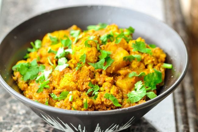 Cauliflower and potato dry curry in a black bowl and garnished with chopped coriander. Bowl is on top of a brown marble window ledge.