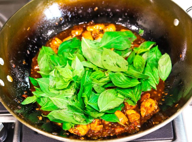 thai sweet basil leaves thrown on top of thai basil chicken that is cooking in a wok on the stovetop