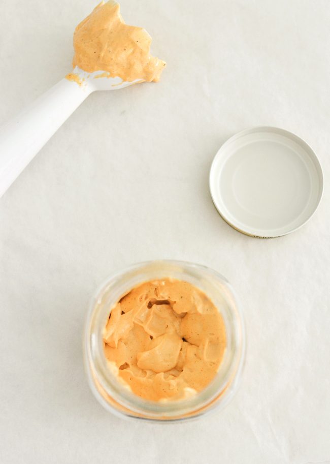 handheld blender's blade covered in spicy mayonnaise, jar with orange mayonnaise, and jar lid on white background.