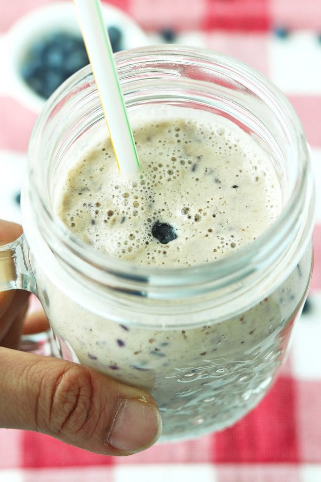 Hand holding up a mason jar mug with a frothing smoothie and a straw, and there's a sinking blueberry at the top of the drink.