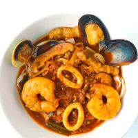 Squid, prawns, mussels, pork, and mushrooms, in a spicy red soup with noodles in a white bowl.
