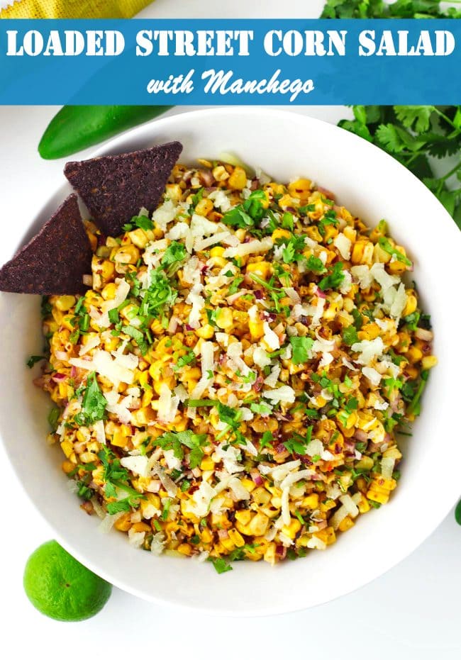 Corn salad in a bowl with two blue corn tortilla chips. Jalapeño, lime, and coriander bunch surrounding bowl.