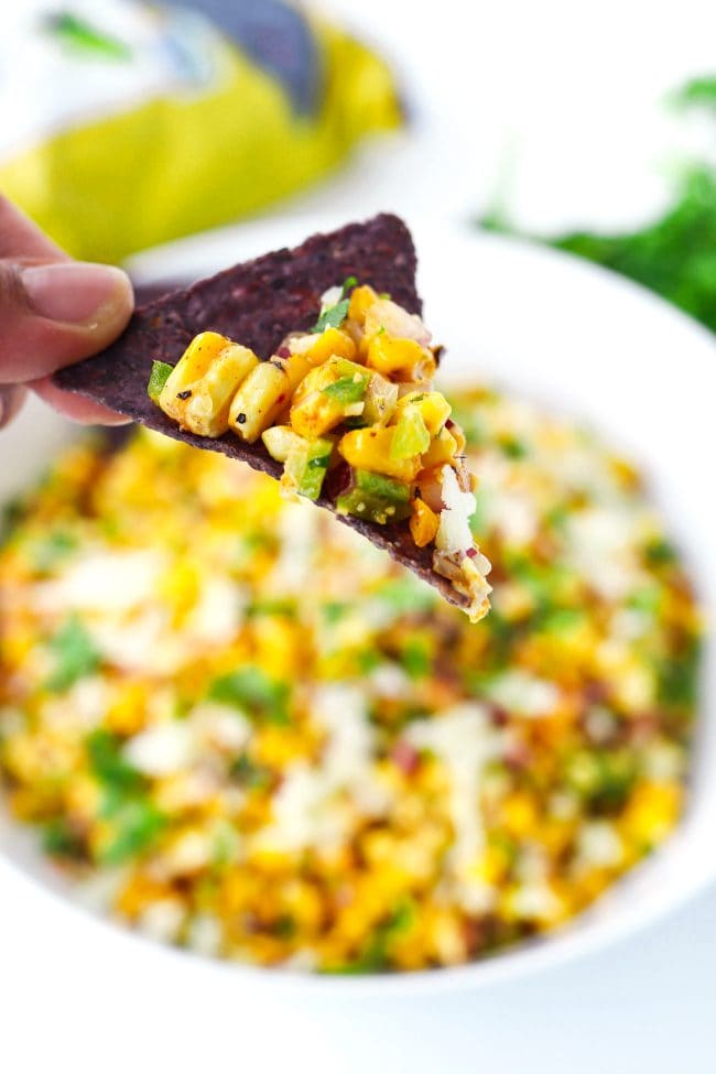 Fingers holding up a blue corn tortilla chip with some corn salad above a bowl filled with corn salad.
