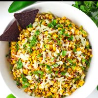 Corn salad garnished with cheese and coriander in a bowl with two blue corn tortilla chips. Jalapeño, lime, and coriander bunch surrounding bowl.