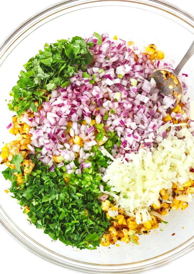 Corn, seasonings, spices, chopped red onion, chopped parsley, chopped coriander, chopped jalapeño, and grated cheese in a large mixing bowl with a silver spoon.