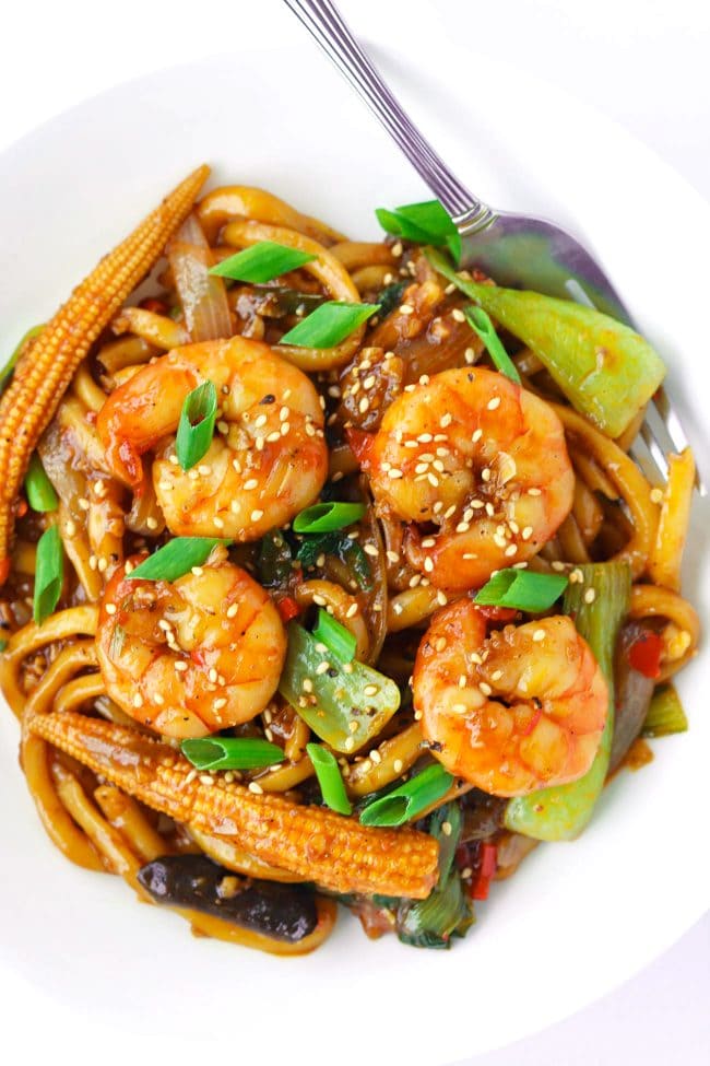 Asian stir-fry noodles with prawns, baby corn, mushrooms, bok choy, and spring onion garnish on a white plate with a fork.