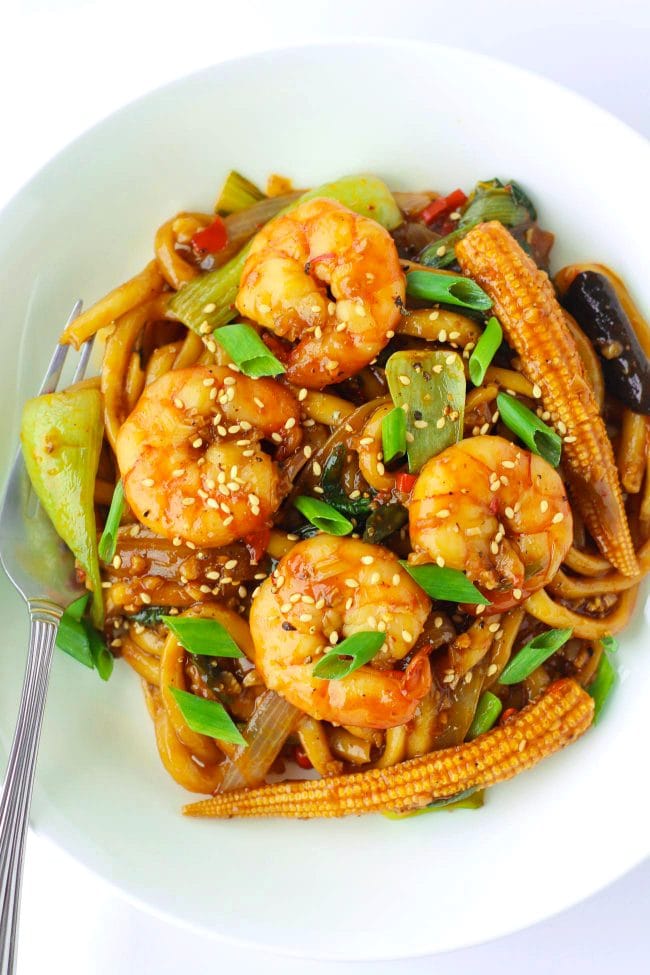 Stir-fry Sichuan Sauce Noodles with Jumbo prawns, baby corn, mushrooms, bok choy, and spring onion garnish on a white plate with a fork.