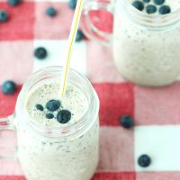 Two mason jar mugs with blueberry and banana smoothie with a straw in each mug. Blueberries scattered around the mug on a red and white checkered napkin.