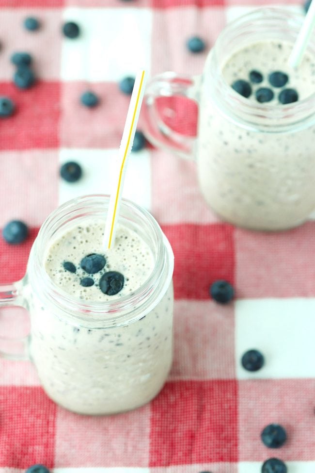 Two mason jar mugs with blueberry banana smoothie with a straw in each mug. Blueberries scattered around the mug on a red and white checkered napkin.