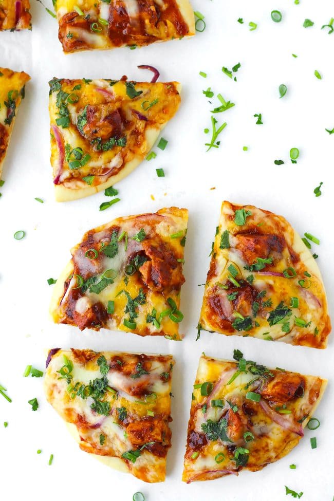 Two diagonally placed Spicy BBQ Chicken Pita Pizzas garnished with spring onion and chopped coriander. Sliced and separated quarters on a white background.