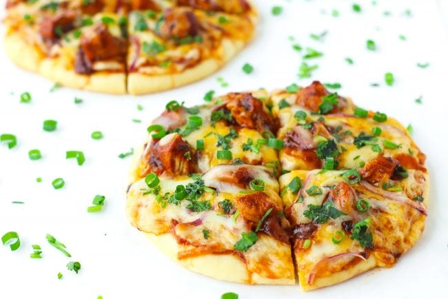 Two diagonally placed sliced Spicy BBQ Chicken Pita Pizzas garnished with spring onion and chopped coriander on top of a white background.