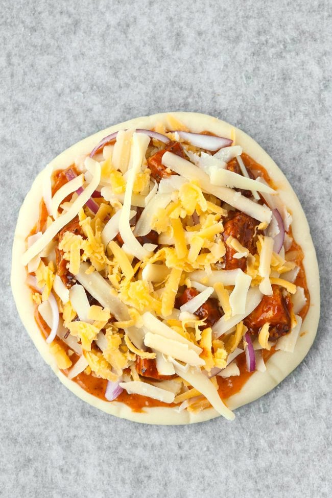 unbaked pita pizza with onion slices, shredded cheese, chicken, and sauce on nonstick cooking paper.
