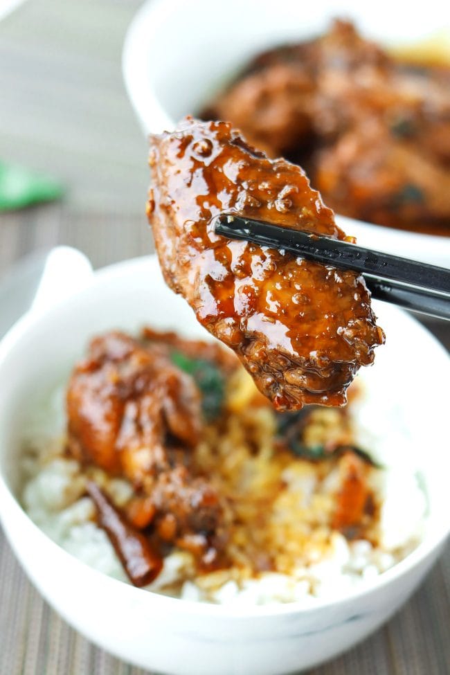 Chopsticks holding up a brown sauce glazed chicken wing above a bowl filled with rice and another chicken wing and sauce.