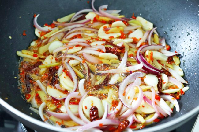 Sliced garlic, ginger, dried red chilies, fresh red chilies, and sliced red onion sizzling in sesame oil in a wok.