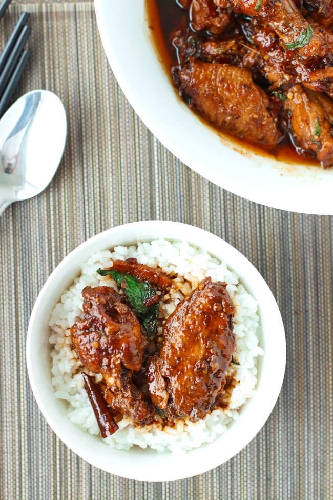 Two pieces of brown sauce glazed chicken wings with sauce on top of steamed white rice in a bowl with a spoon. Dish of San Bei Ji above the bowl. Spoon and chopsticks to the side of the bowl.