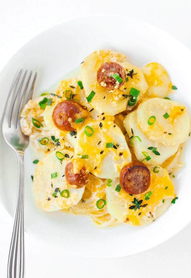 Sliced sausage and cheesy potato rounds garnished with spring onion and fresh thyme in a white round plate with a fork to the side of the potatoes.