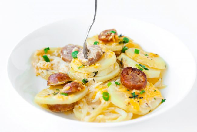 Sliced sausage and cheesy potato rounds garnished with spring onion and fresh thyme in a white round plate. A fork is sticking out of a sausage slice and three potato rounds in the center of the plate.