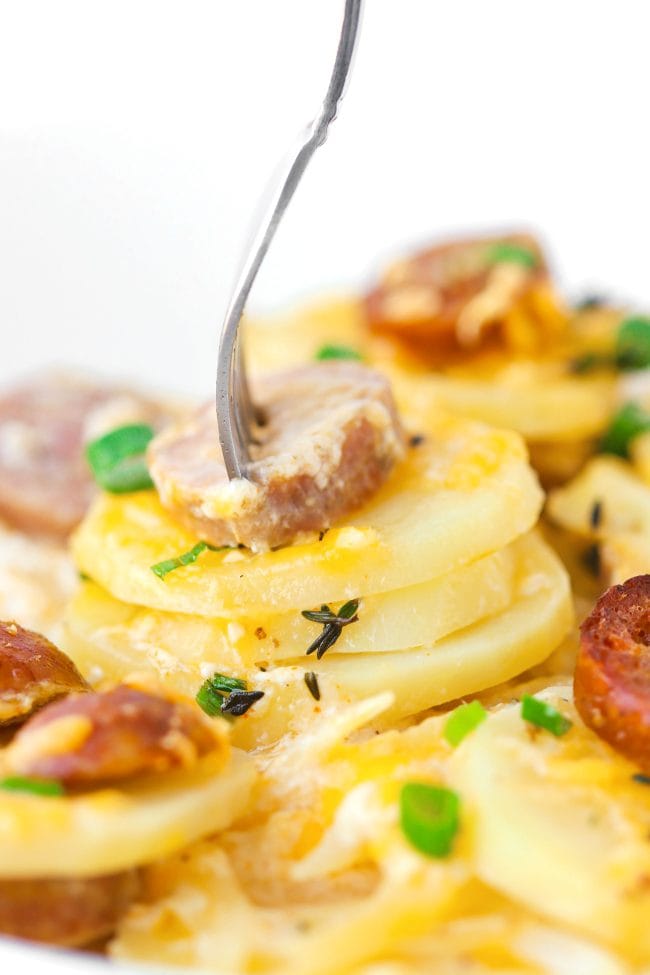 Sliced sausage and cheesy potato rounds garnished with spring onion and fresh thyme in a plate with a fork sticking out of a sausage slice and three potato rounds.