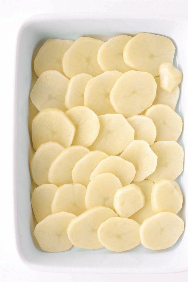 Thinly sliced potato rounds lined up in an even layer at the bottom of a large rectangular white baking dish.