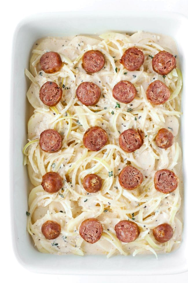Thinly sliced potato, spicy garlic cream sauce with sliced onion, and sliced sausage rounds in a large rectangular white baking dish.