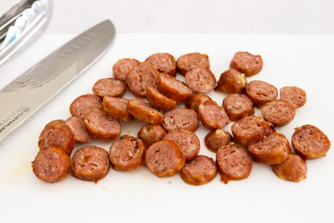 Sliced rounds of cooked sausage on a chopping board with a knife and tongs.