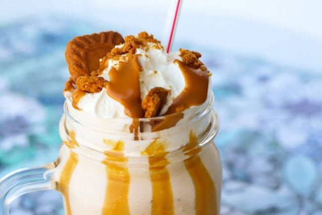 Cookie butter milkshake in a mason jar glass with a straw, whipped cream, crumbled cookies, and melted cookie butter drizzle.