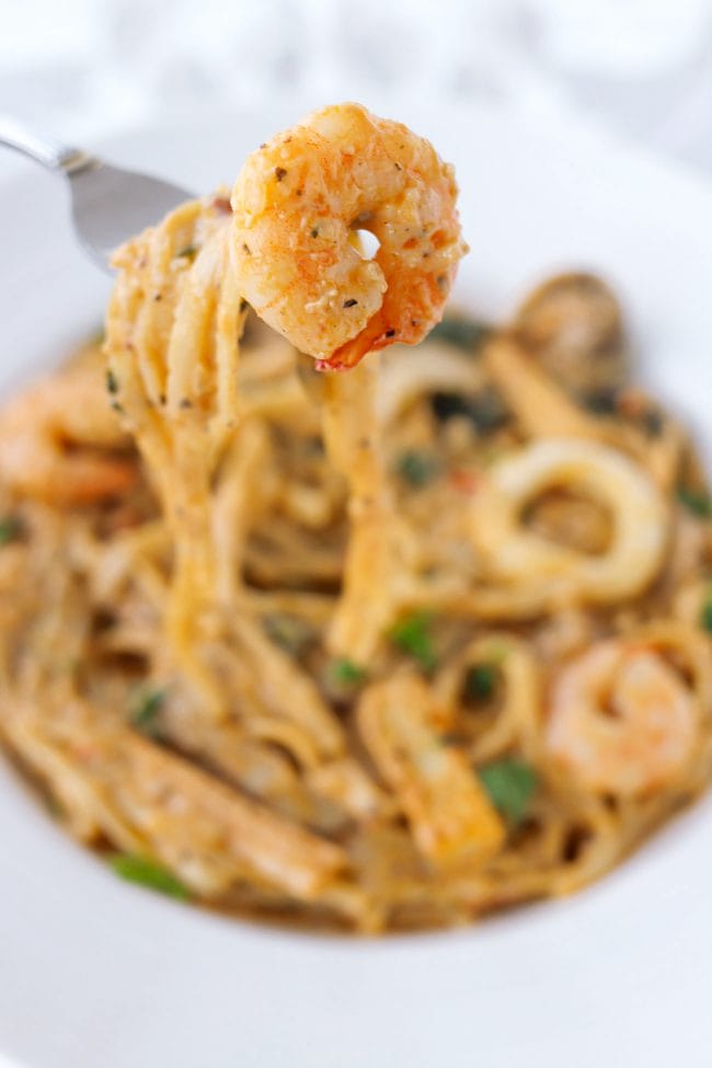 Hand holding up a fork with a piece of shrimp and pasta above plate with pasta and seafood.