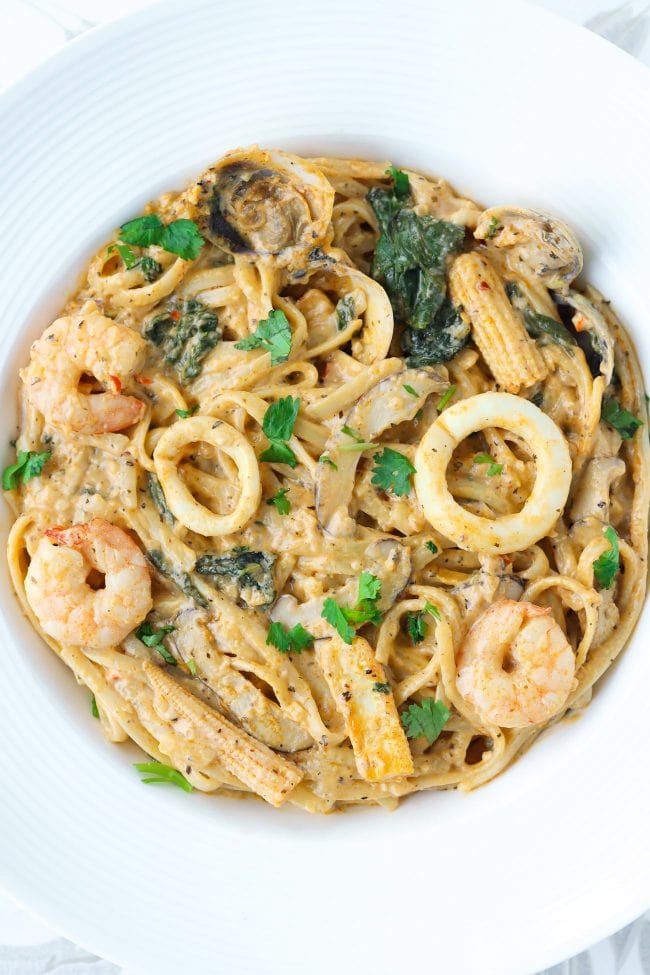 Pasta in tom yum cream sauce with seafood, baby spinach, baby corn, and sliced mushrooms in a white round plate.