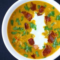 Pumpkin Chicken Soup garnished with chopped coriander, yogurt, and crispy bacon pieces in a white round bowl.