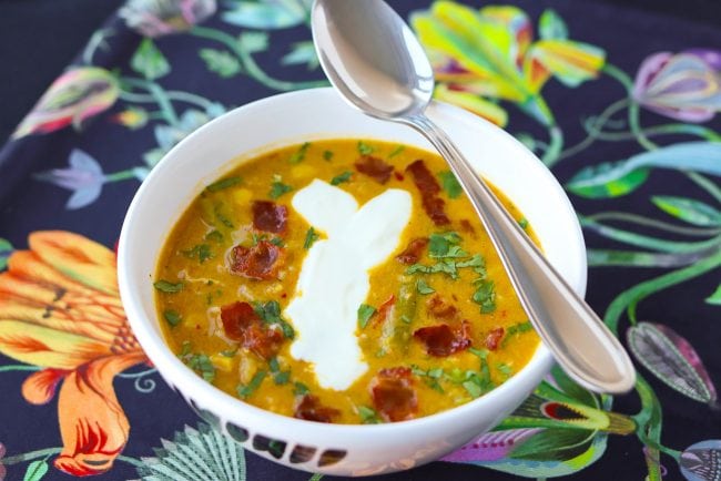 Soup in bowl topped with crispy bacon pieces, yogurt, chopped coriander and a spoon on top of bowl.