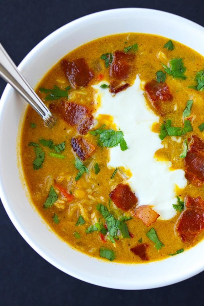 Soup in bowl with crispy bacon pieces, yogurt, chopped coriander and spoon inside bowl.