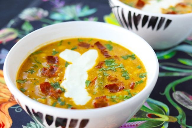 Two diagonally placed bowls of Pumpkin Chicken Soup garnished with chopped coriander, yogurt, and crispy bacon pieces.