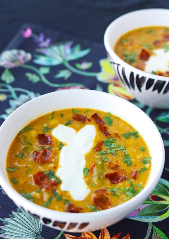 Two diagonally placed bowls of soup garnished with chopped coriander, yogurt, and crispy bacon pieces.