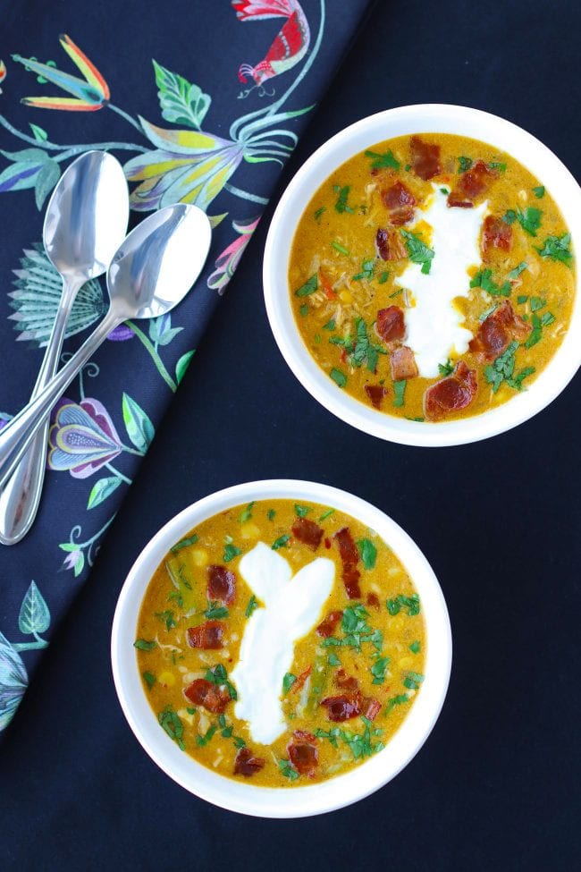 Two diagonally placed bowls of soup garnished with chopped coriander, yogurt, and crispy bacon pieces. Two spoons on top of a blue floral printed napkin on the side.