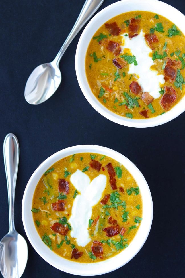Two diagonally placed bowls of Pumpkin Chicken Soup garnished with chopped coriander, yogurt, and crispy bacon pieces. Two spoons on the side of bowls.