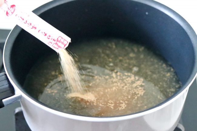 Hand pouring a sachet of dashi powder into a pot of simmering water on the stovetop.