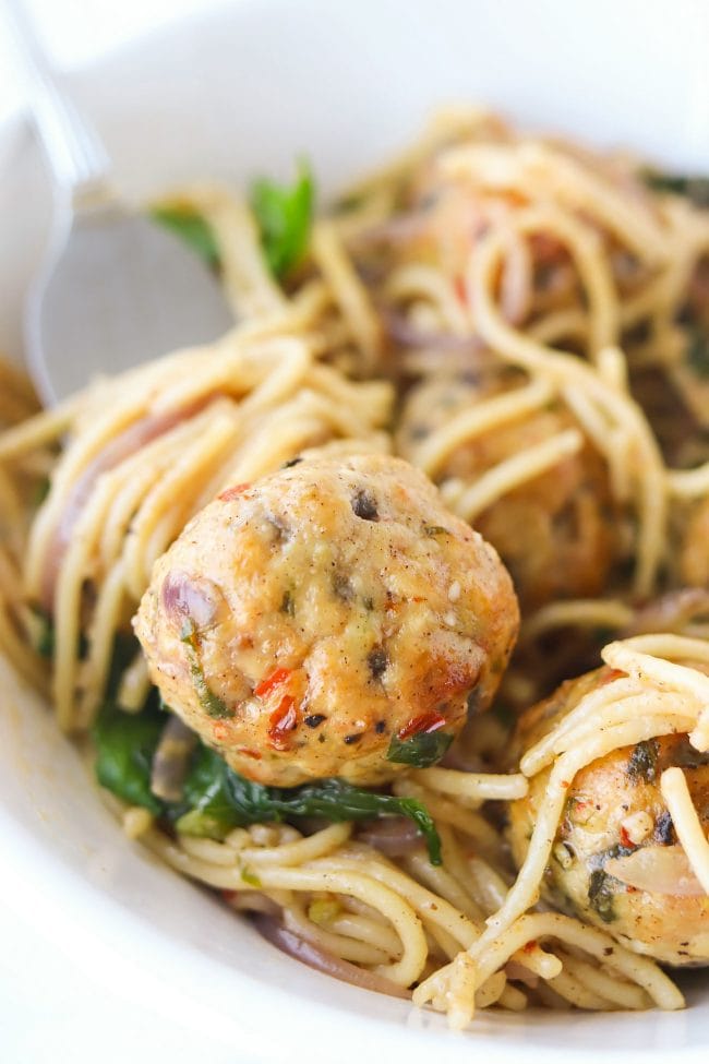 Fork twirled around capellini pasta and stabbed into a chicken meatball in a bowl of pasta and chicken meatballs with baby kale and red onion slices.