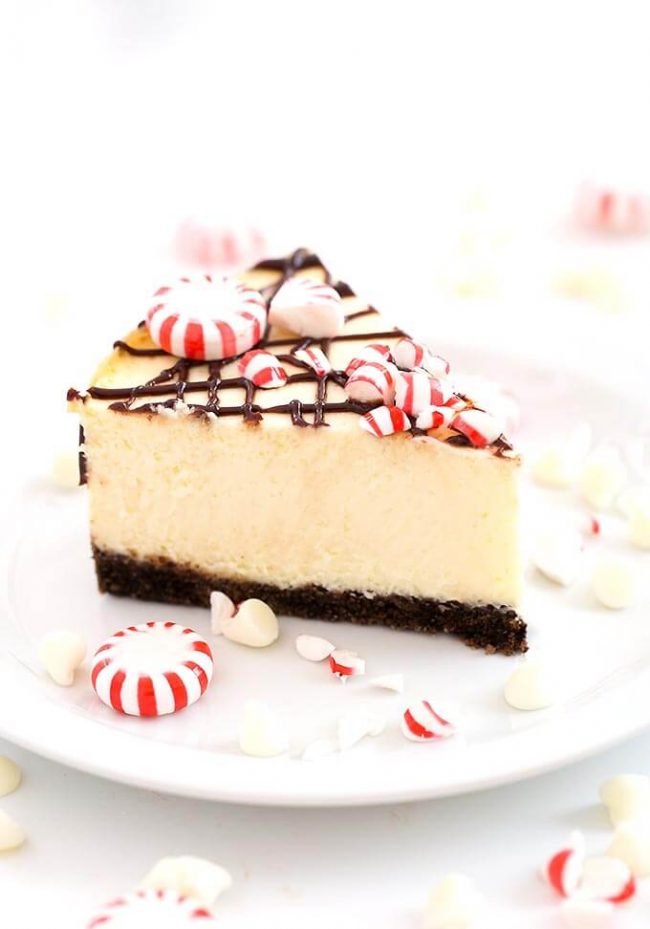 Peppermint and White Chocolate Cheesecake slice with a chocolate cookie crust on a plate. Cake is topped with peppermint candy and chocolate ganache drizzle.