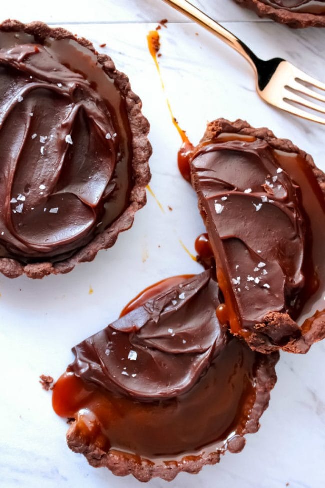 Salted Caramel Chocolate Tarts topped with chocolate ganache and sea salt flakes on a marble background. One of the broken tarts has gooey caramel oozing out.