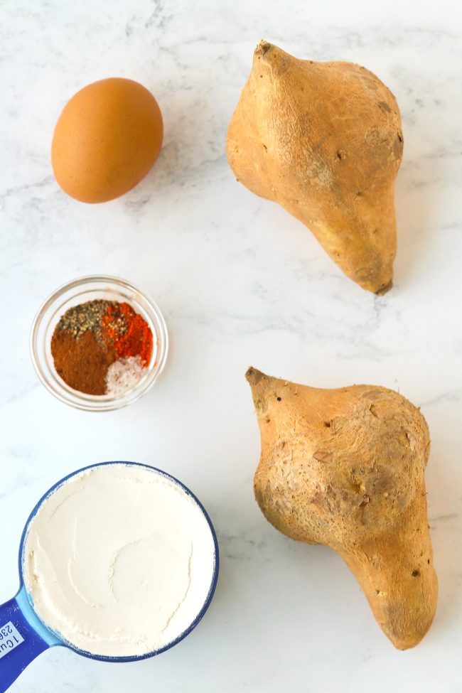 Two sweet potatoes, a measuring cup filled with flour, seasonings and spices in a small glass bowl, and an egg.
