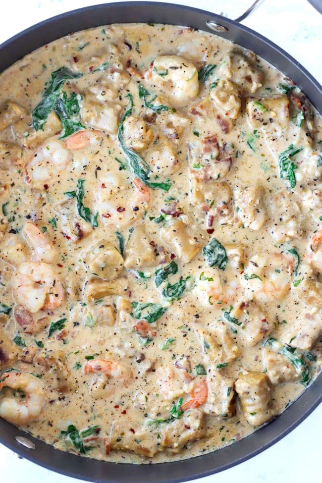 A deep large black sauté pan with sweet potato gnocchi, crispy bacon, shrimp, baby kale, and parsley in a spicy white wine garlic cream sauce.