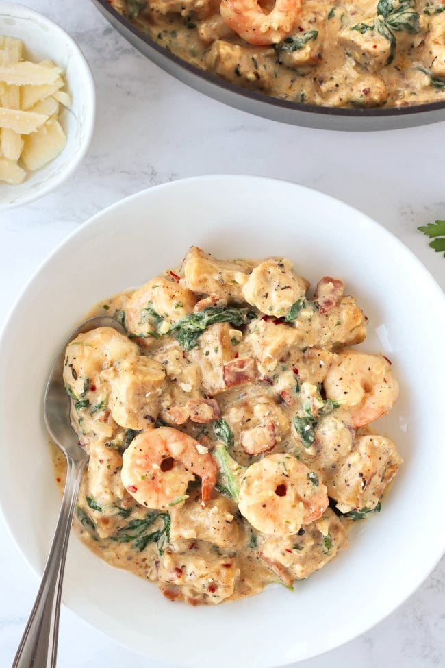 A round white plate with a spoon and creamy sweet potato gnocchi with shrimp, baby kale, and bacon. A pan with the creamy sweet potato gnocchi and a small bowl with Parmigiano Reggiano cheese shavings in the back.