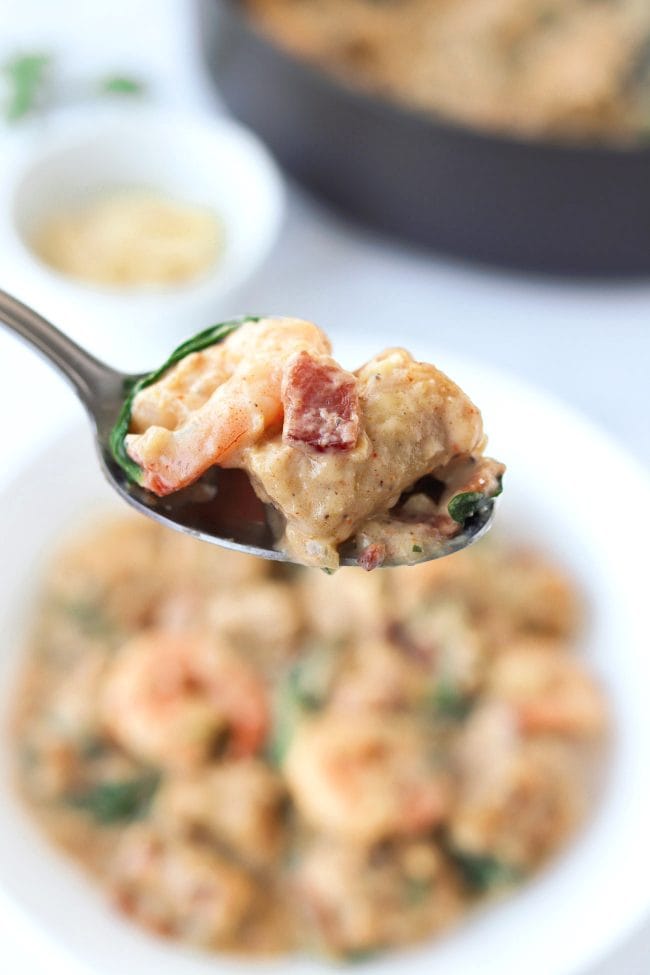 A spoon holding up a piece of creamy sweet potato gnocchi with bacon, shrimp, and baby kale over a plate with creamy sweet potato gnocchi.