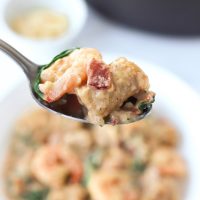 A spoon holding up a piece of creamy sweet potato gnocchi with bacon, shrimp, and baby kale over a plate with creamy sweet potato gnocchi.