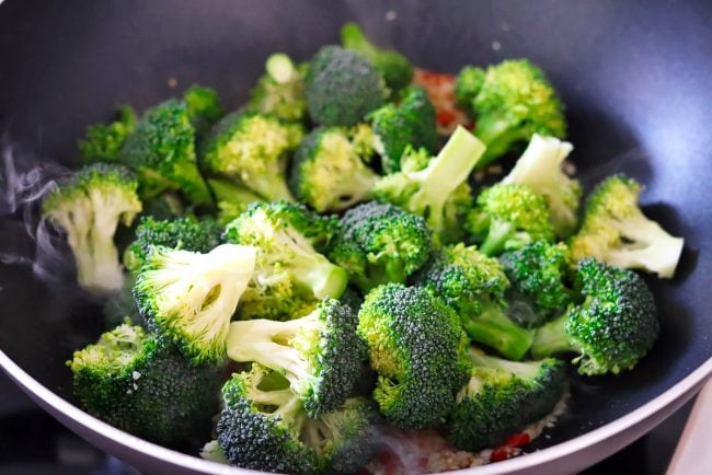 Broccoli florets with garlic and chopped red chiles in a wok with smoke coming out.