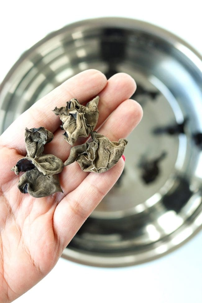 Hand holding up four pieces of dried cloud ear mushrooms above a stainless steel bowl with dried wood ear mushrooms soaking in water