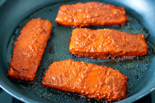 Four salmon fillets coated in an orange-red spicy paste marinade pan-frying skin side down in a large nonstick skillet on the stovetop.