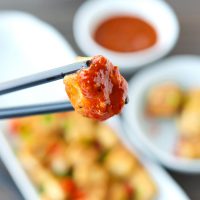 Black chopsticks holding up a fried tofu cube that's dipped in a spicy orange sauce above a plate of pan-fried tofu, bowl with sauce, and a small plate.