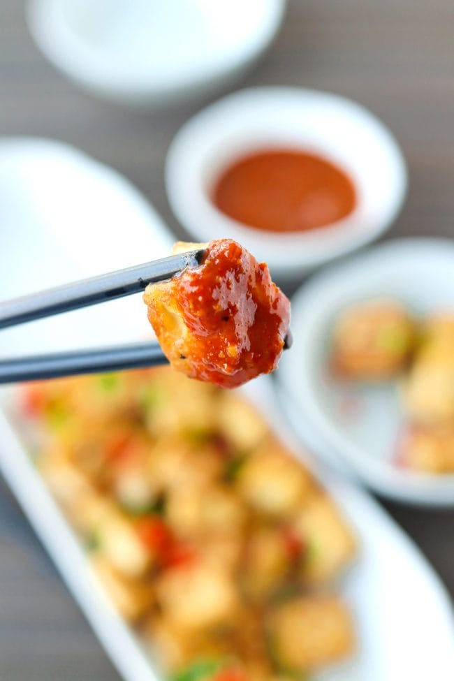 Black chopsticks holding up a fried tofu cube that's been dipped in a spicy orange sauce above a plate with pan-fried tofu sprinkled with chopped spring onion, chopped red chili, and toasted white sesame seeds.